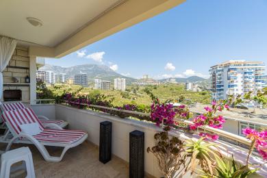 Alanya ejendom balkon med loungestole, lyse blomster og fantastisk udsigt over bjergene, der skaber en afslappende atmosfære.