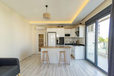 Kuchyně v nemovitosti Alanya s bílými skříňkami, ostrůvkem s barovými židlemi, světlou dřevěnou podlahou a světlou, vzdušnou atmosférou.