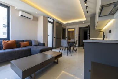 La propriété d'Alanya dispose d'un salon moderne avec un canapé gris élégant, un coin repas et un éclairage encastré lumineux, créant une atmosphère chaleureuse.