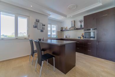 Alanijos virtuvė su tamsaus medžio spintelėmis, sala su sėdimomis vietomis, plytelių backsplash ir ryškia natūralia šviesa.