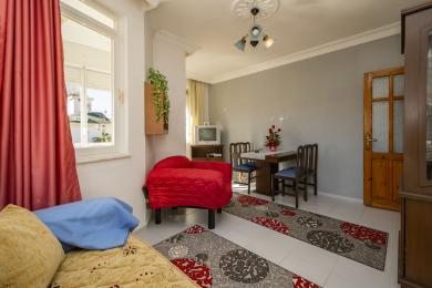 Obývací pokoj apartmánu v Alanyi s jasným oknem, červenými akcenty, dlaždicovou podlahou a útulnou, zabydlenou atmosférou.
