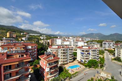 Alanya mulkining ko'rinishi, unda ko'p qavatli uylar, basseyn va yorqin, quyoshli osmon ostidagi tog'lar namoyish etilgan.