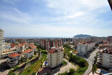 Alanya mulkidan ko'rinish: O'rta yer dengizi va tog'lar uzoqda ko'rinadigan yorug', quyoshli shahar manzarasi.