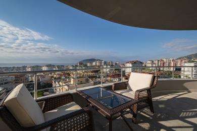Alanya shahridagi balkon, to‘qilgan mebellar va shisha stol bilan yorug‘ kunduzda ajoyib shahar va dengiz manzaralarini taqdim etadi.