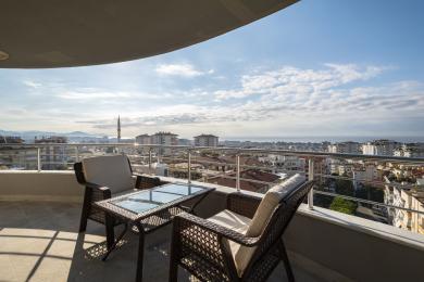 Alanya mulkidagi balkon, to‘qilgan mebel, shisha stol va shahar va dengizning panoramik ko‘rinishlari bilan.