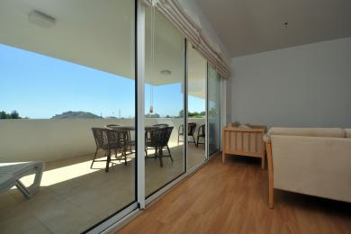 Sala de estar luminosa em Alanya com piso de madeira e portas de vidro deslizantes que se abrem para uma varanda ensolarada com assentos ao ar livre.