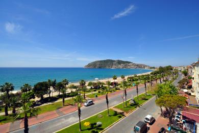 Vista del balcón de la propiedad de Alanya que muestra la playa, las palmeras y el cielo azul claro en un día soleado.