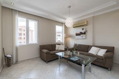 Sala de estar do apartamento Alanya com sofás castanhos, mesa de vidro, pisos de cerâmica e luz natural brilhante.