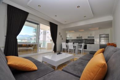 Alanya'da satılık mülk: Açık mutfaklı, balkon erişimli, gri kanepeli ve parlak, doğal ışıklı modern oturma odası.