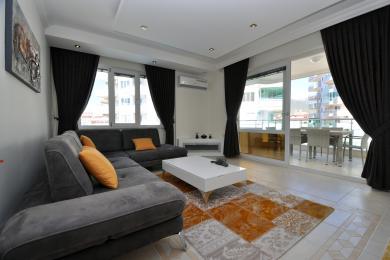 Gri bir kanepe, balkon erişimi, koyu renk perdeler ve modern beyaz mobilyalar içeren aydınlık Alanya oturma odası.