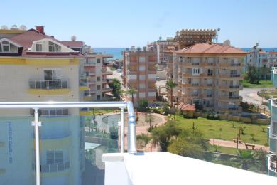 Alanya mulkidagi balkon manzarasi obodonlashtirilgan hovliga, yaqin atrofdagi binolarga va yorqin osmon ostidagi O'rta er dengiziga qaraydi.