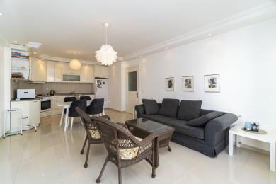 Šviesus Alanya buto svetainės kambarys su atvira virtuve, plytelių grindimis, pintais krėslais ir patogia pilka sofa.