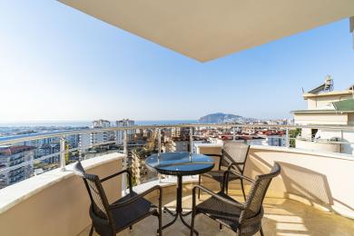 Balcone di Alanya con sedie in vimini e tavolo di vetro, che offre viste panoramiche sulla città e sul mare in piena luce solare.